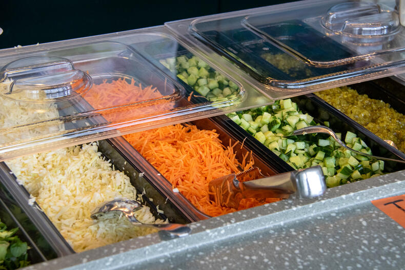 Ruokalan linjasto, jossa kapeissa astioissa kaaliraastetta, porkkanaraastetta ja kurkkua kuutioina.