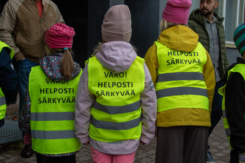 Kolme lasta seisoo rivissä, kaikilla on keltaiset heijastinliivit joiden selässä lukee: Varo! Helposti särkyvää