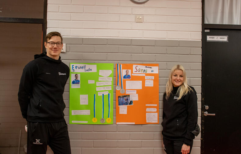 Hymyilevät, mustiin pukeutuneet Eemeli Salin ja Sanni Nieminen seisovat seinän edessä, heidän väliinsä on kiinnitetty värikkäät pahvit, joissa on tietoa pelaajista.