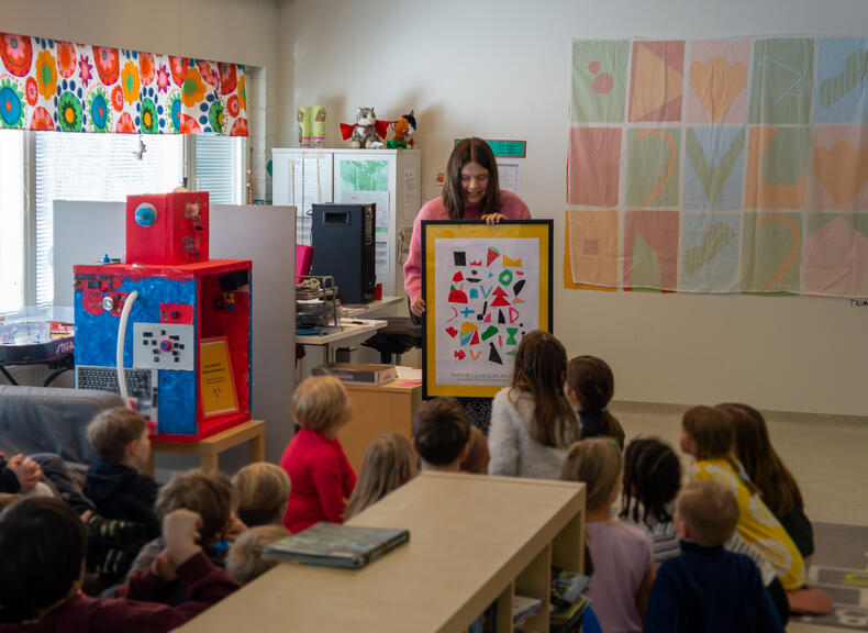 Heidi Häyry esittelee lapsille kehystettyä taulua, jossa on erilaisia muotoja, apip-toiminnan lasten kehittämät salakielen aakkoset.