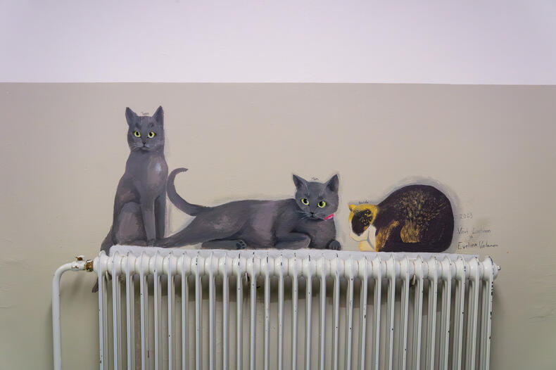Seinällä olevan patterin päälle on maalattu kolme kissaa; yksi istuu, toinen makaa ja kolmas on kerällä.