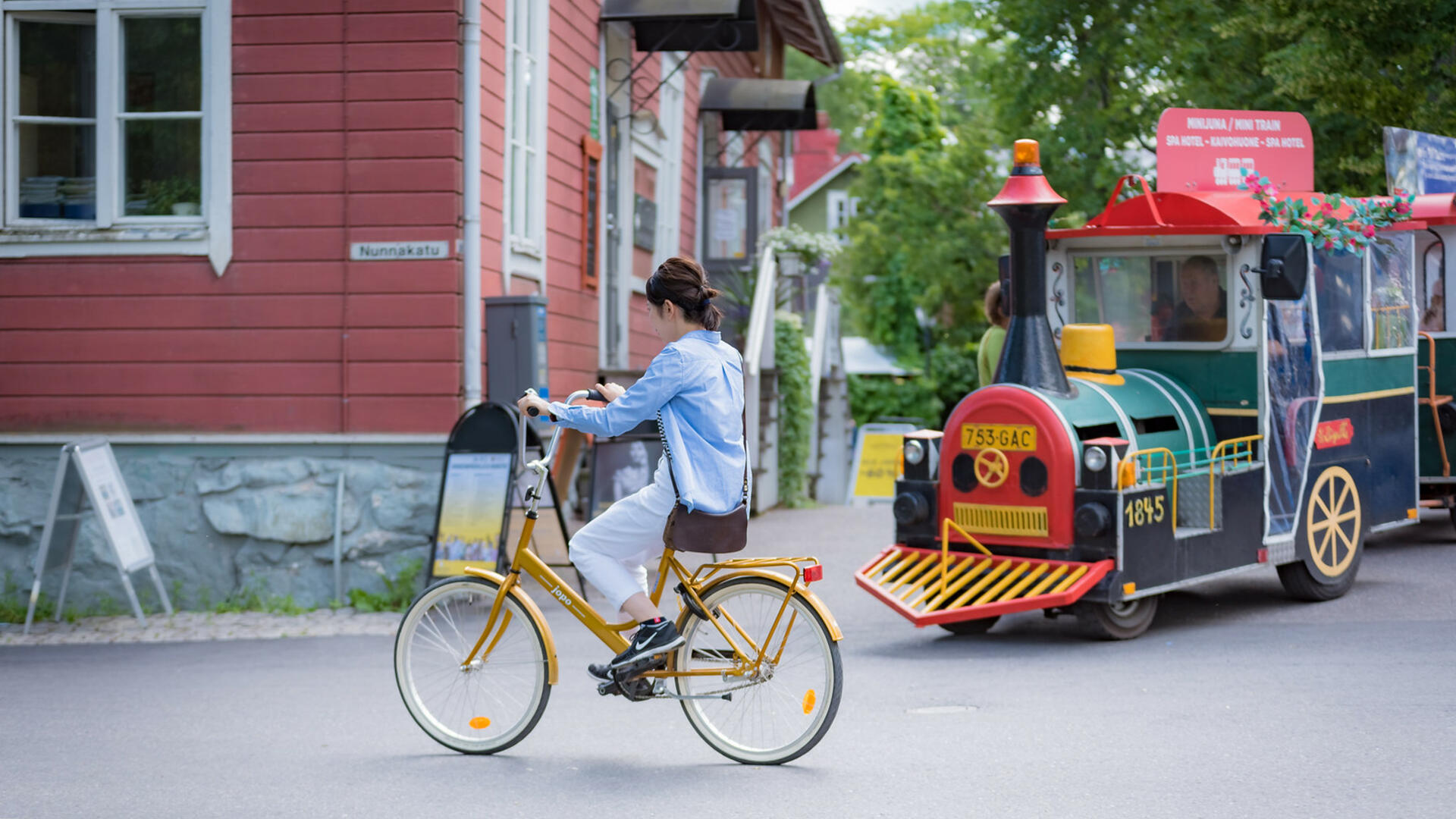 Nuori nainen pyöräilee jopolla Naantalin Vanhassakaupungissa, taustalla kaupunkijuna sekä punainen puutalo.