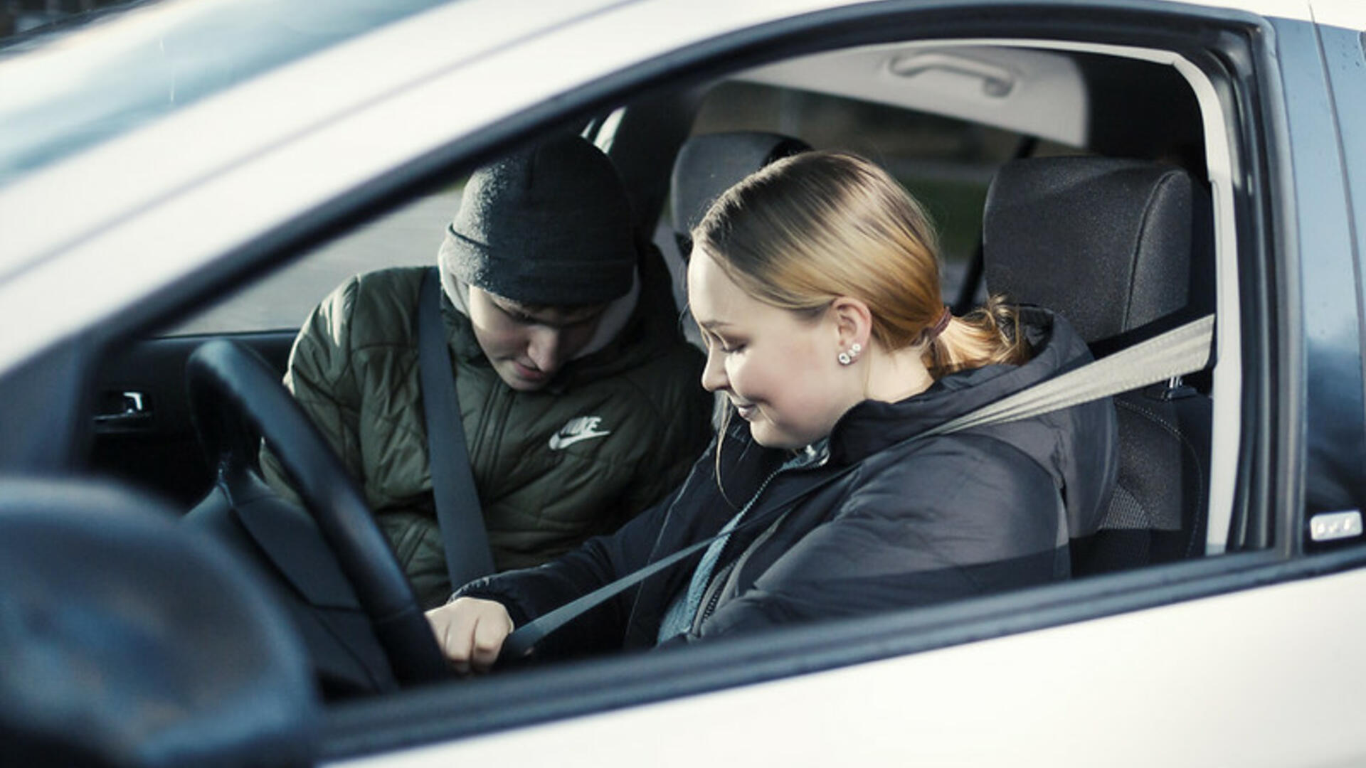 Nuori nainen istuu henkilöauton ratissa ja etupenkillä istuu nuori mies, molemmat kiinnittävät turvavöitä.
