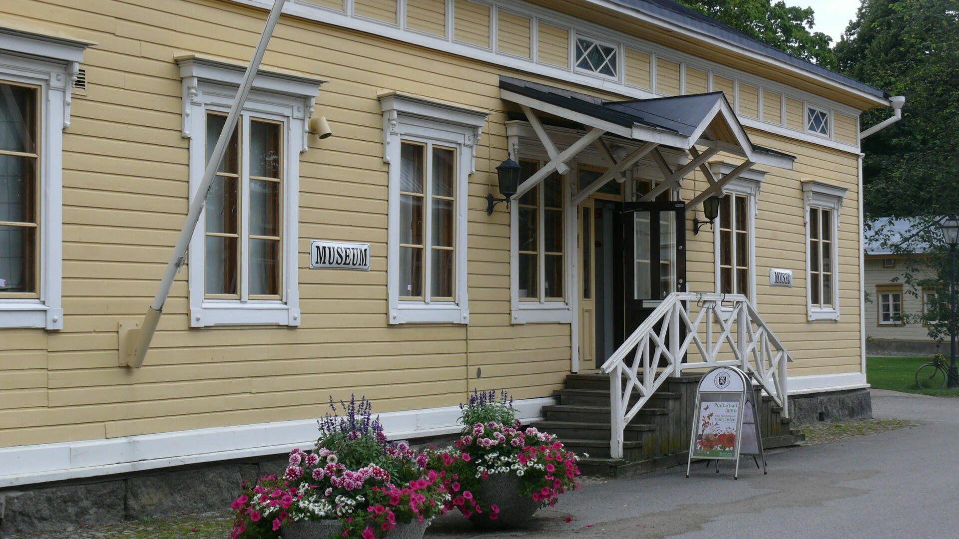 Vanha puurakennus jonka edustalla kukkaistutuksia, museon ovi avoinna 