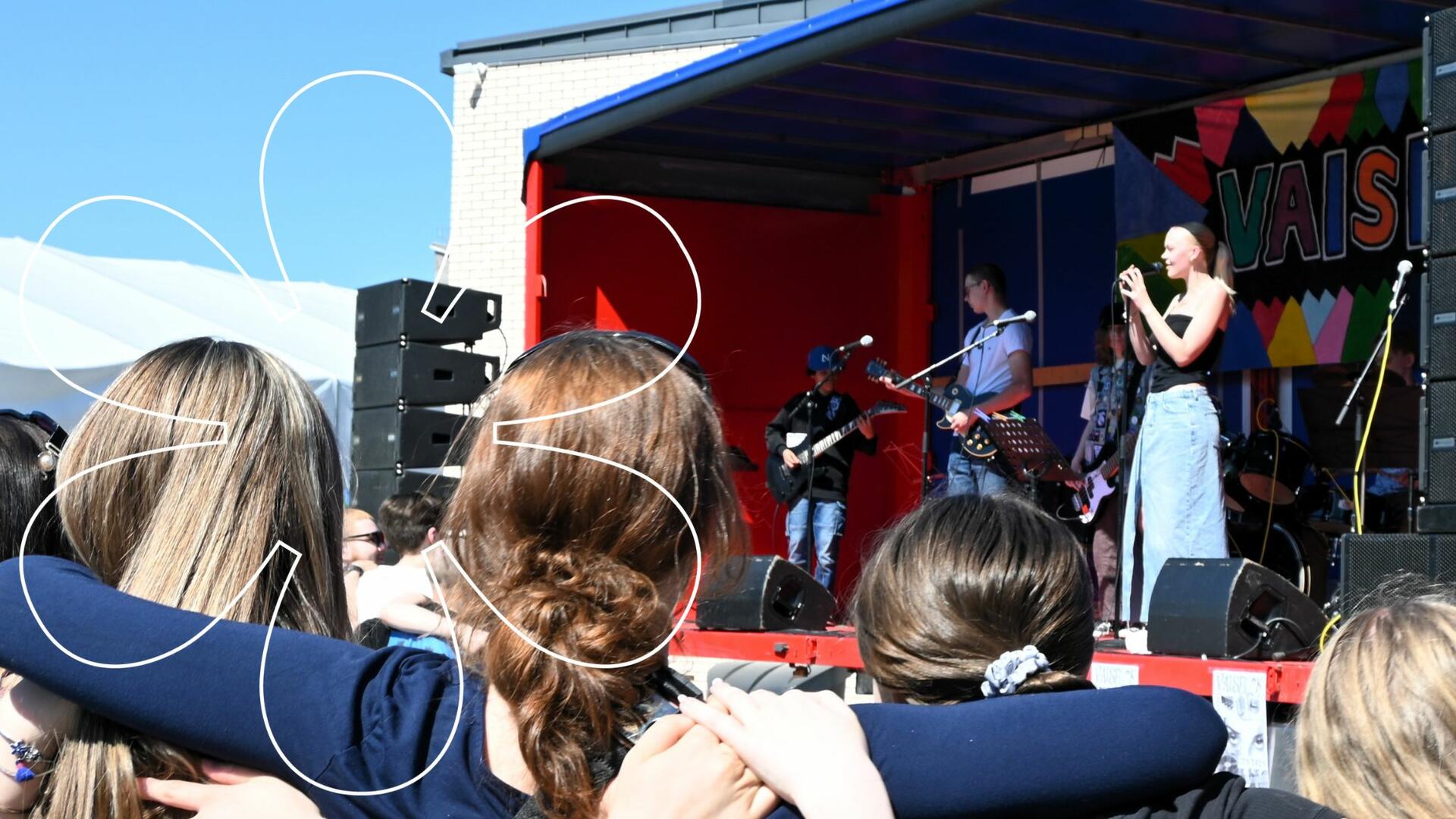 Vaisrockin lavalla Maksuhäiriömerkintä-bändi esiintymässä, etualalla yleisöä. Onnellinen aurinko -elementti.