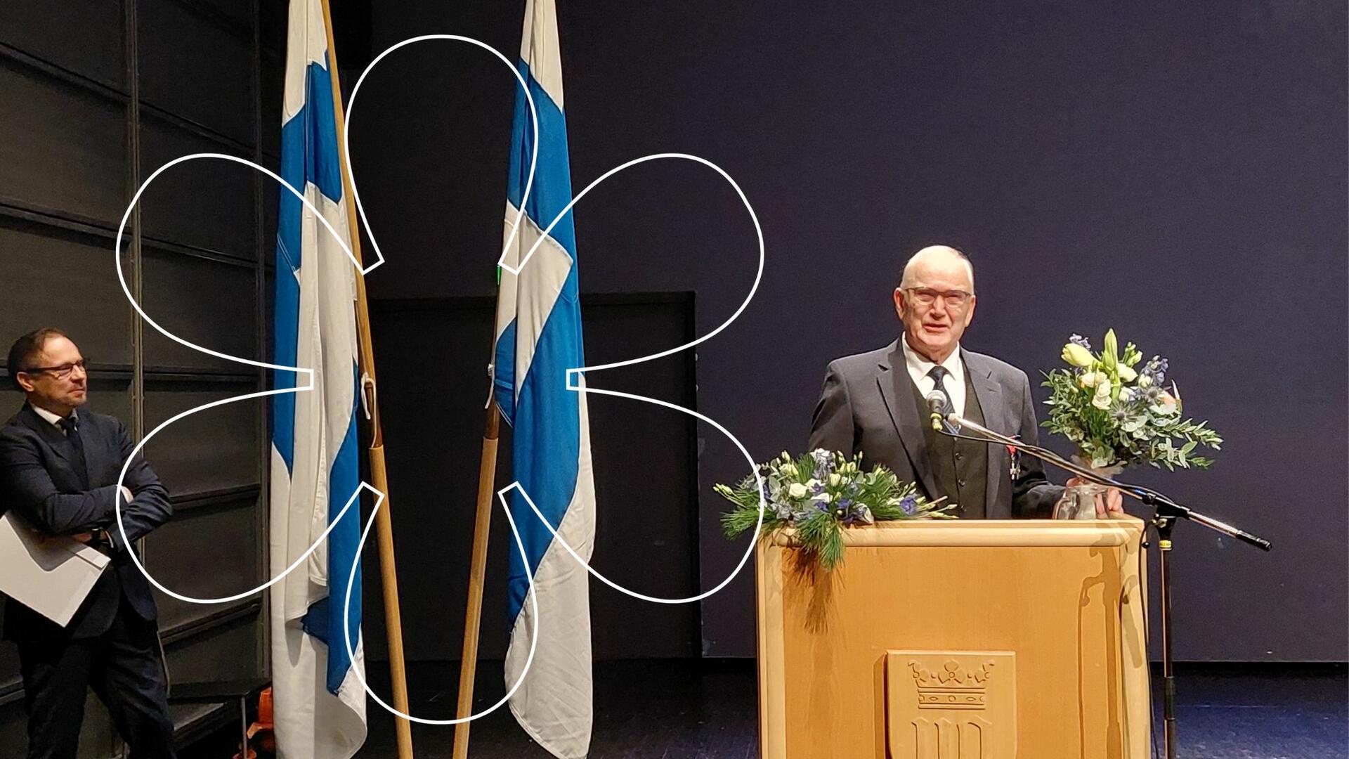 Raimo Laine pitää puhujanpöntössä puhetta, Suomen liput kohoavat vieressä. Sivussa seuraa Sami Rantanen. Kuvan päällä Onnellinen aurinko -elementti.