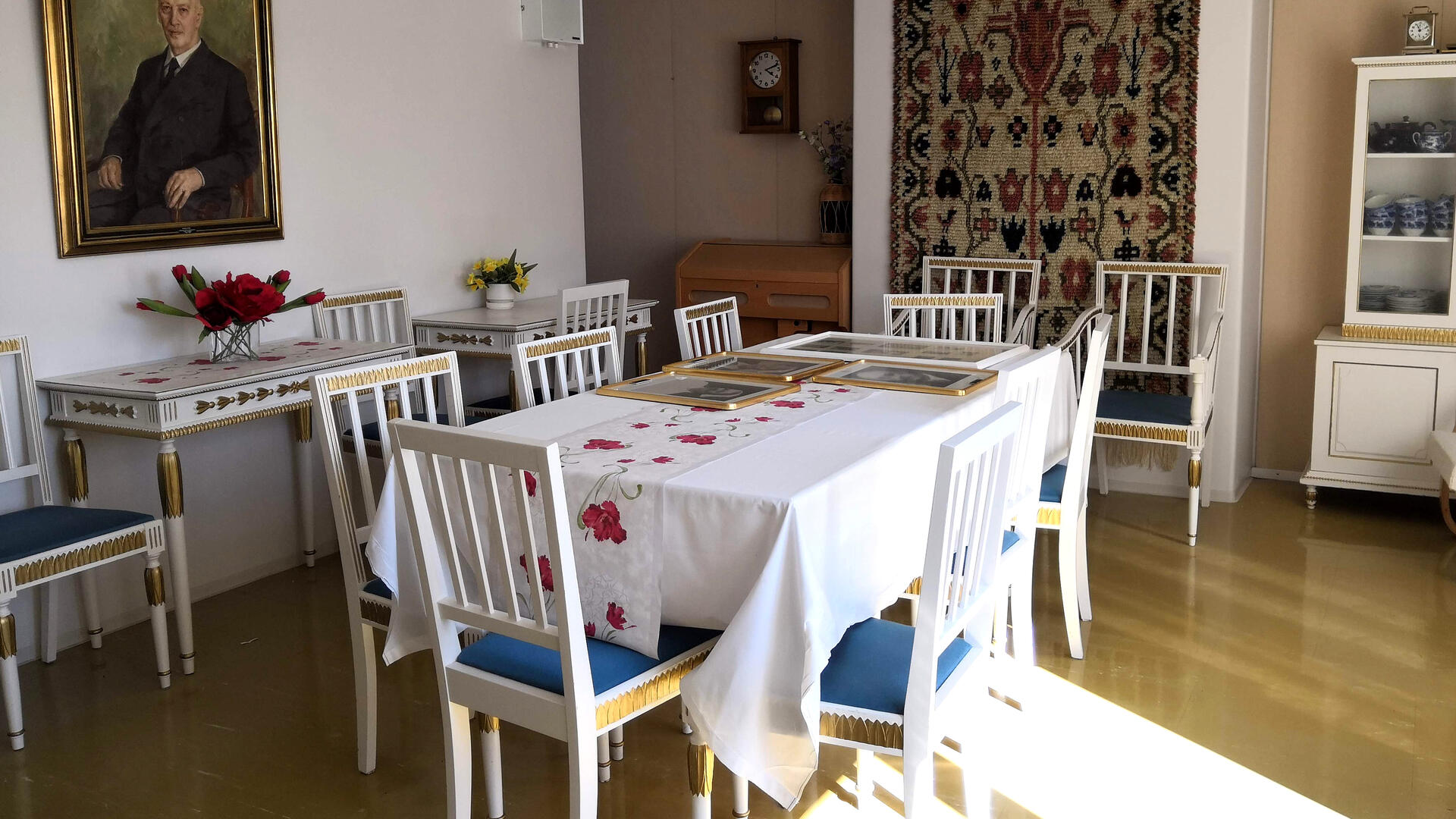 huoneessa valkoinen ruokapöytä tuoleineen, taustalla apupöydät, seinällä kehystetty maalaus ja ryijy
