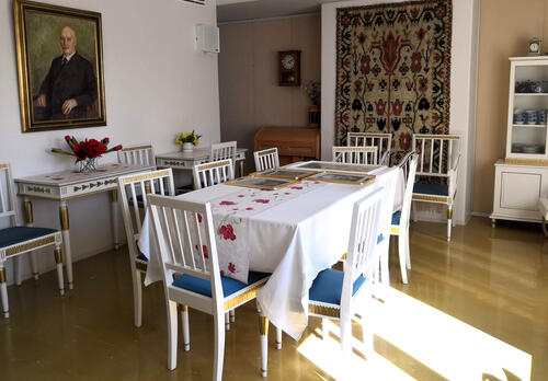 huoneessa valkoinen ruokapöytä tuoleineen, taustalla apupöydät, seinällä kehystetty maalaus ja ryijy